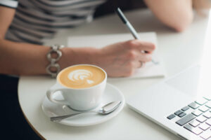 Ilustracja do tekstu Skuteczny tekst reklamowy. Kobieta siedzi przy biurku i pisze odręcznie w notesie. Obok stoi laptop i filiżanka kawy.